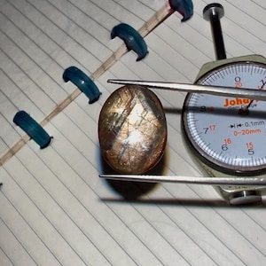 May include: An oval-shaped gemstone with brown and gold hues, held by tweezers. A dial gauge, measuring in millimetres, is positioned nearby. The gem rests on lined paper, suggesting a gemological setting.