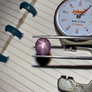 May include: A round, purplish-pink gemstone with a star-like pattern, held by a gem gauge. The gauge displays measurements in millimeters. The gemstone is positioned on a lined notepad, with several blue binder clips visible.