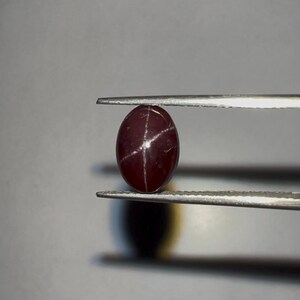 May include: A polished, oval-shaped, dark red gemstone held by tweezers. The gemstone has a star-shaped pattern of light lines across its surface. The background is a neutral grey.