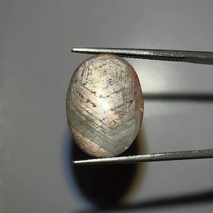 Pode incluir: Uma pedra preciosa ovalada segurada por pinças. A gema tem uma mistura de cores, incluindo cinza, marrom e uma pitada de rosa. A superfície exibe um padrão único, possivelmente um efeito de estrela, com um design geométrico.