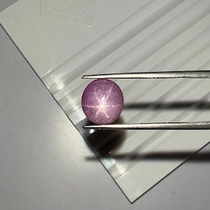 May include: A round, polished, pink gemstone with a star-like pattern, held by tweezers. The gem is set against a white background with a diamond-shaped design. The gem's surface reflects light, creating a star effect.
