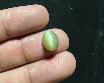 Puede incluir: Una piedra preciosa ovalada de color verde con un efecto chatoyante. La piedra se sostiene en una mano.