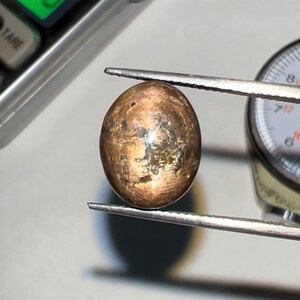 May include: A brown and gold oval-shaped gemstone with a metallic sheen, being measured with a gauge.