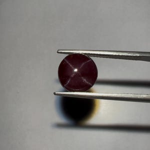May include: A round, dark red gemstone with a star pattern, held by silver tweezers. The gem has a polished surface and a star effect. The background is a neutral grey.