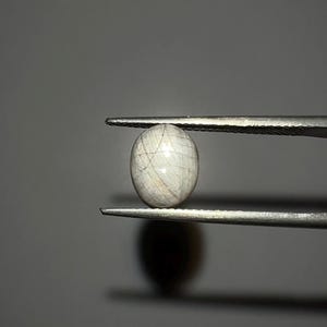 May include: An oval gemstone, held by tweezers, with a pale, translucent appearance. Fine, hair-like inclusions are visible within the gem. The background is a plain, neutral grey.