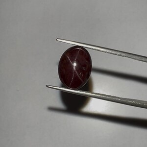 May include: A polished, oval-shaped, dark red gemstone held by tweezers. The gem has a star-like pattern of light reflecting across its surface. The background is a neutral grey.