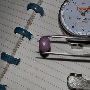 May include: A pinkish-purple star sapphire gemstone held by a jewelry caliper. The gemstone has a six-rayed star pattern. A dial gauge is in the background, along with several blue ring sizers on a lined paper.