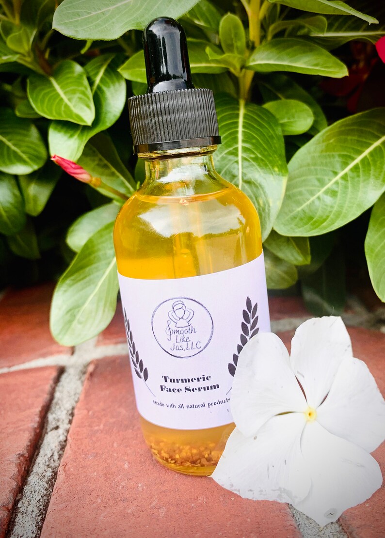 K&ouml;nnte beinhalten: Eine durchsichtige Glasflasche mit einer Tropfpipette, gef&uuml;llt mit einer gelben Fl&uuml;ssigkeit. Die Flasche hat ein wei&szlig;es Etikett mit schwarzem Text, auf dem "Smooth Like Jas, LLC" und "Turmeric Face Serum" steht, darunter die Worte "Made with all natural products". Die Flasche steht auf einer Ziegelstein-Oberfl&auml;che mit einer wei&szlig;en Blume.