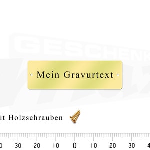 May include: A gold-colored metal plaque with the text "Mein Gravurtext" and two small holes for mounting. The plaque is shown on a white background with a ruler in the background.