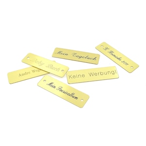May include: Six gold-colored metal tags with different text engraved on them. The text includes "Baby Buch", "Mein Tagebuch", "Keine Werbung", "Andre Wege", "Mein Poesiealbum", and "H. Heimicke 2011".