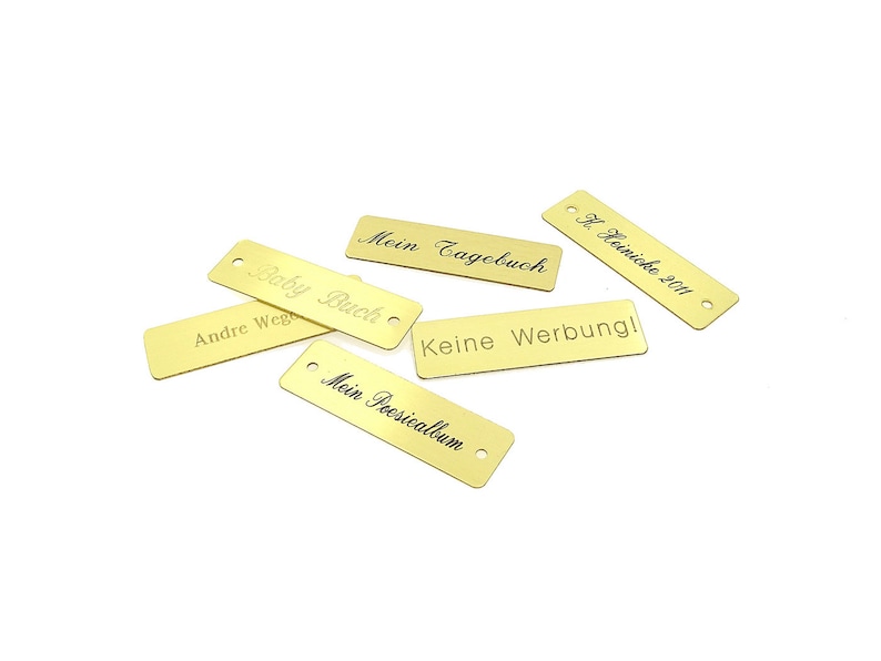 May include: Six gold-colored metal tags with different text engraved on them. The text includes "Baby Buch", "Mein Tagebuch", "Keine Werbung", "Andre Wege", "Mein Poesiealbum", and "H. Heimicke 2011".