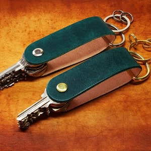 May include: Two green leather keychains with silver and gold key rings. The keychains are made of dark green leather with a light pink backing. The key rings are attached to a bundle of keys.