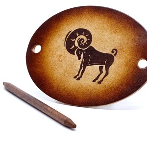 May include: An oval hair stick made of brown leather with a cutout of the zodiac sign Aries, a ram with horns, in the centre. The hair stick is on a white background.