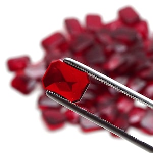 Peut inclure: Une seule pierre précieuse rouge facetée, brillante, tenue par des pinces sur un fond de nombreuses pierres précieuses rouges similaires.