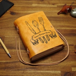 Könnte beinhalten: Ein braunes, ledergebundenes Tagebuch mit einem Rezept-Thema. Der Umschlag zeigt eine Zeichnung einer Gabel, eines Messers und eines Löffels mit dem Text "Annas Rezepte Buch".