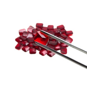 May include: A single, bright red, faceted gemstone is held by silver tweezers, with a pile of similar gems in the background. The gems are a deep red colour and have a square shape. The background is plain white.