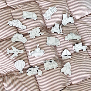 May include: A collection of unpainted wooden vehicle cutouts, including a car, airplane, submarine, and rocket. The vehicles are arranged on a striped, reddish-brown fabric background. The collection includes a variety of transportation types.