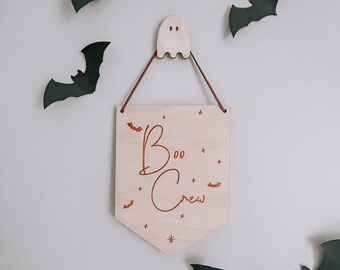 May include: A wooden sign with the text "Boo Crew" in a cursive font. The sign is hanging from a string and has a ghost-shaped hook. There are black bats cutouts on a white wall around the sign.