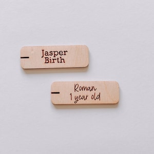May include: Two rectangular wooden plaques with dark brown text. One reads "Jasper Birth" and the other reads "Roman 1 year old". The plaques are light brown and have a small black line on the left side.