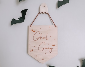 May include: A wooden sign with the words "Ghoul Gang" in a script font, hanging from a ghost-shaped hanger. The sign is decorated with small bats and stars. Black bat cutouts are affixed to the white wall around the sign.