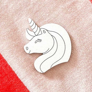 May include: A white wooden cutout of a unicorn head with a single horn and closed eyes. The cutout is on a pink and red striped towel.