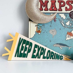 May include: A wooden sign with the text "KEEP EXPLORING" in green letters on a white background. The sign is shaped like a pennant and has yellow accents. A gold glitter crescent moon and a book titled "MAPS" are in the background.