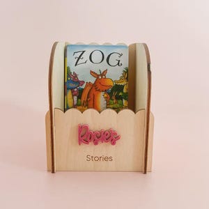 May include: A light-coloured wooden book holder with a scalloped top edge. The front features the pink script text 'Rosie's' above the word 'Stories' in a simple font. Inside the holder sits a Zog book with a colourful dragon illustration.