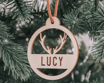 May include: A wooden Christmas ornament with the name "LUCY" cut out in a circular design. The ornament features a deer antler silhouette and is suspended by a brown leather cord. The ornament hangs on a green Christmas tree.