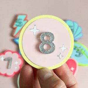 May include: A pink and yellow circle with a silver glitter number 8 in the centre. The circle has a yellow border and two white glitter stars on either side of the number.