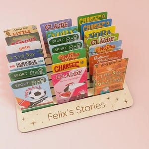 May include: A light-coloured wooden display stand with horizontal slots holding multiple rectangular Yoto audio cards. The front of the stand is engraved with the text "Felix's Stories" and small star icons. The cards feature various colourful illustrations and titles.