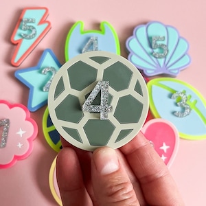 May include: A set of nine colorful felt patches with different shapes and numbers. The patches are decorated with glitter and feature designs like a lightning bolt, a star, a seashell, a football, and a heart. The number four is on a green soccer ball patch.
