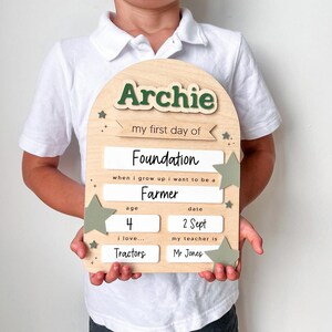 May include: An arched wooden sign with the name Archie in dark green letters. It features text fields for school details including my first day of Foundation, when i grow up i want to be a Farmer, age 4, date 2 Sept, i love Tractors, and my teacher is Mr Jones, decorated with small sage green stars.