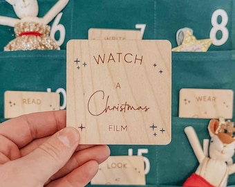 May include: A wooden square with the words "WATCH A Christmas FILM" in brown script. The square is held in a hand, and is part of a Christmas advent calendar. The background is a teal green fabric with small pockets.