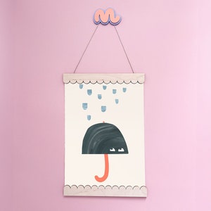 May include: A white framed print with a black umbrella with orange handle and blue raindrops falling on it. The print is hanging on a pink wall with a pink and blue cloud hanging above it.