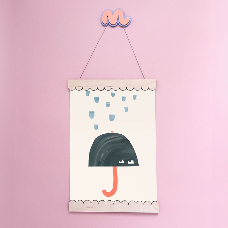 May include: A white framed print with a black umbrella with orange handle and blue raindrops falling on it. The print is hanging on a pink wall with a pink and blue cloud hanging above it.