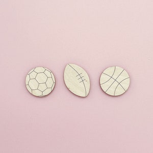 May include: Three wooden cutouts of sports balls on a pink background. The cutouts are of a football, a rugby ball, and a basketball.