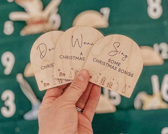 May include: Three wooden Christmas-themed tags with text. The tags are held in a hand against a green background. The text on the tags reads "Decorate Christmas", "Wear A Christmas", and "Sing Some Christmas Songs". Each tag has a small illustration of houses and trees.