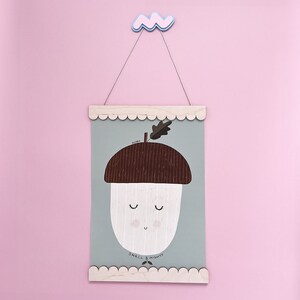 May include: A light blue and white illustrated print of an acorn with a smiling face. The print is framed with a light wood frame and has a scalloped edge. The print reads "ACORN SMALL & MIGHTY!"