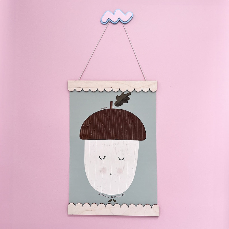 May include: A light blue and white illustrated print of an acorn with a smiling face. The print is framed with a light wood frame and has a scalloped edge. The print reads "ACORN SMALL & MIGHTY!"