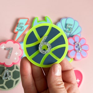 May include: A set of colourful felt patches with numbers and glitter. The patches are shaped like a lightning bolt, a shell, a flower, a turtle, a heart, and a basketball. The basketball patch has a black and green design with the number 6 in the centre.