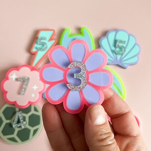May include: A set of colourful felt patches with numbers on them. The patches are shaped like a lightning bolt, a flower, a seashell, a turtle, and a cloud. The numbers are 3, 4, 5, 7, and 5.