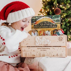 May include: A wooden crate labelled "Sofia" filled with Christmas gifts and decorations. The crate is decorated with festive illustrations and the text "A special Christmas delivery Love Santa x". A Christmas tree and a book are in the background.