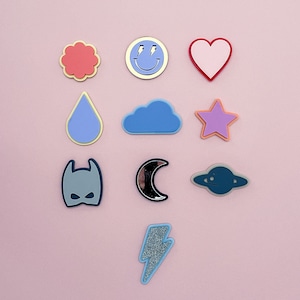 May include: A set of ten colorful iron-on patches featuring various shapes and designs, including a smiley face, a heart, a flower, a raindrop, a cloud, a star, a bat, a crescent moon, a spaceship, and a lightning bolt.