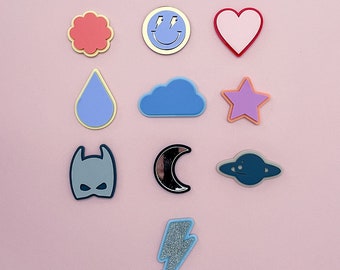 May include: A set of ten colorful iron-on patches featuring various shapes and designs, including a smiley face, a heart, a flower, a raindrop, a cloud, a star, a bat, a crescent moon, a spaceship, and a lightning bolt.