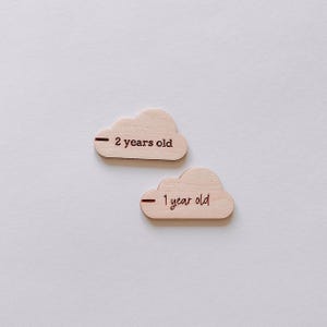 May include: Two wooden cloud-shaped age markers. One marker reads "2 years old" and the other reads "1 year old". The text is in a dark brown color. The clouds are light brown and sit on a white background.