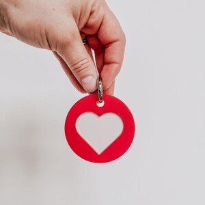 May include: A red heart-shaped tag with a white heart cutout. The tag has a silver ring for attaching to a keychain or bag.