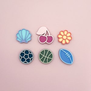 May include: Six colorful, flat, wooden shapes on a pink background. The shapes include a seashell, a cherry, a flower, a soccer ball, a basketball, and a football.