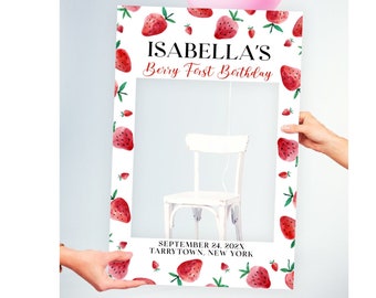 Puede incluir: Un accesorio de fotomatón blanco con un patrón de fresas rojas y verdes. El texto "Isabella's Berry First Birthday" está en la parte superior del marco. La fecha "September 24, 202X" y la ubicación "Tarrytown, New York" están en la parte inferior del marco.