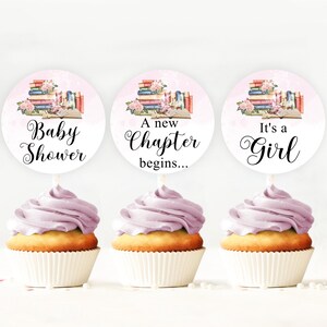 Puede incluir: Tres cupcakes con glaseado morado y adornos redondos. Los adornos muestran una pila de libros con flores y el texto "Baby Shower", "A new Chapter begins...", y "It's a Girl".