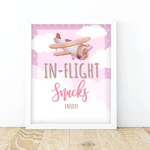 May include: A pink and white printable sign with a cartoon airplane flying in the clouds. The sign says "In-Flight Snacks Enjoy!"